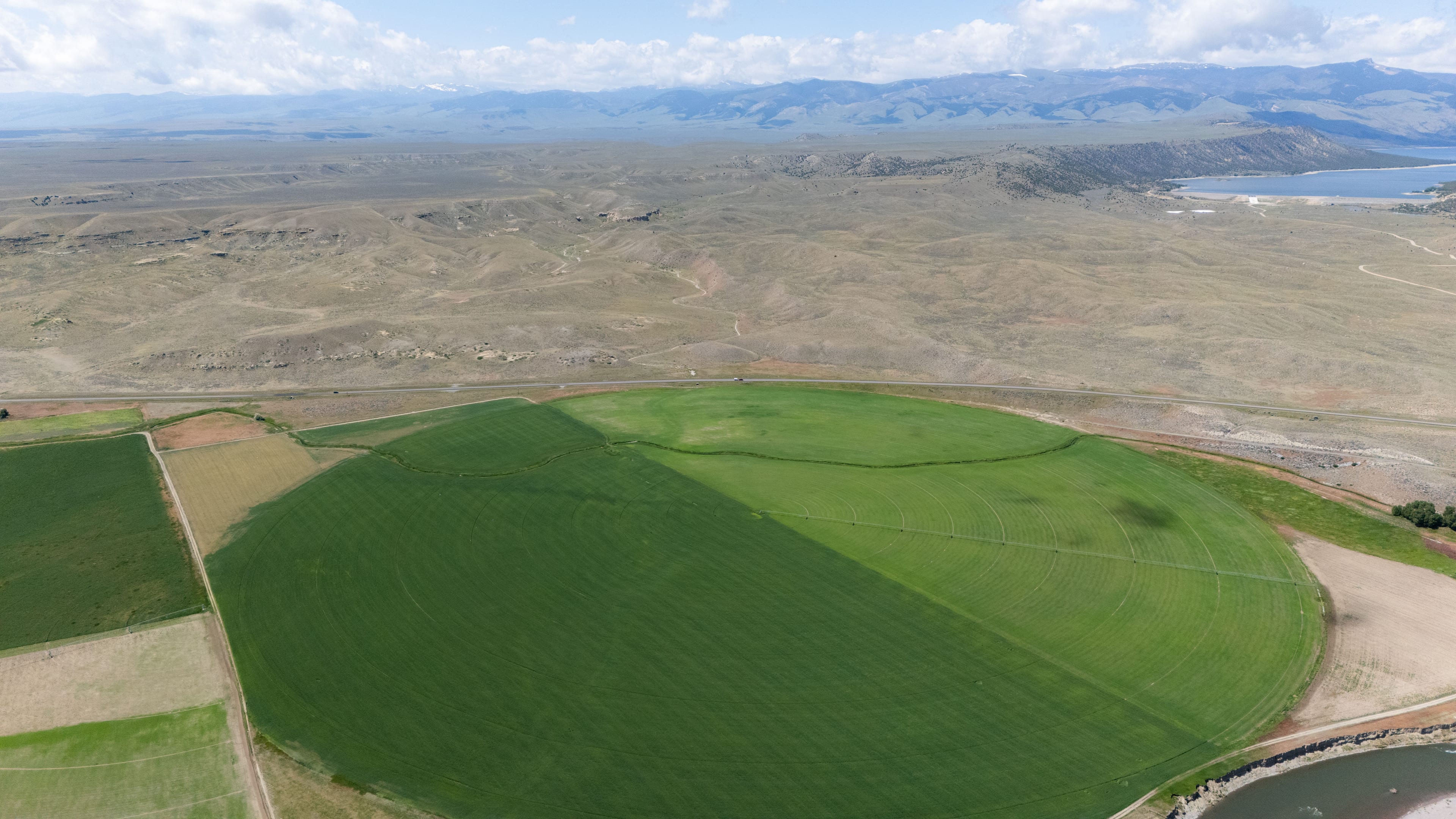 Aerial view of a large green center-pivot circle with smaller fields beside it, dry hills, a reservoir and mountains beyond, under scattered clouds.